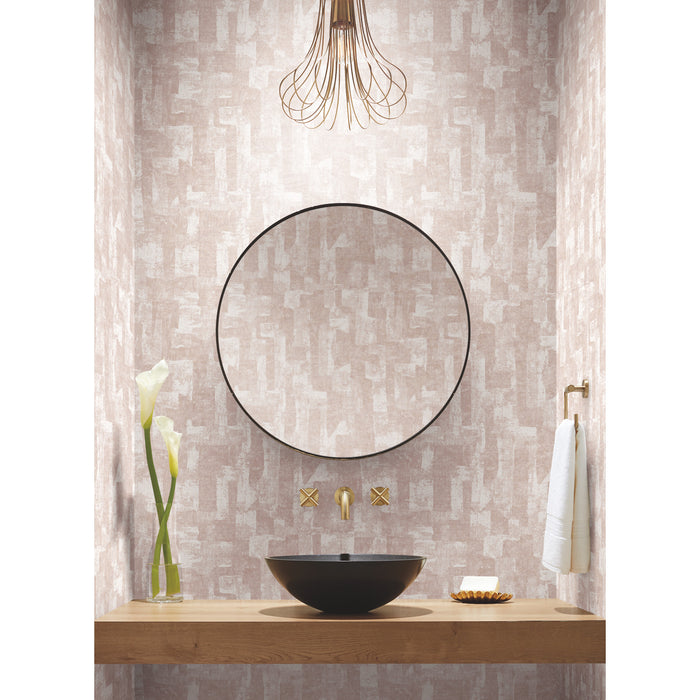 A modern workspace with a laptop, potted plants, an hourglass, a phone, a notebook with a pen, and a cup and saucer on a wooden table. The background features softly blurred blush tones and Nikki Chu Capetown Peel and Stick Wallpaper from WallPops with geometric design, enhanced by warm lights that create a cozy, inviting atmosphere.