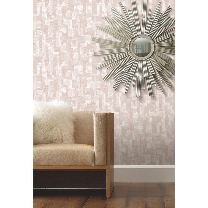 A modern workspace with a laptop, potted plants, an hourglass, a phone, a notebook with a pen, and a cup and saucer on a wooden table. The background features softly blurred blush tones and Nikki Chu Capetown Peel and Stick Wallpaper from WallPops with geometric design, enhanced by warm lights that create a cozy, inviting atmosphere.