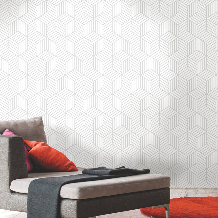 A modern room featuring a gray chaise lounge with orange and red pillows, a dark gray throw, and a vibrant orange circular rug. The wall is adorned with WallPops Striped Hexagon Peel and Stick Wallpaper showcasing a white geometric pattern of thin gray stripes. The overall setting is minimal and stylish.