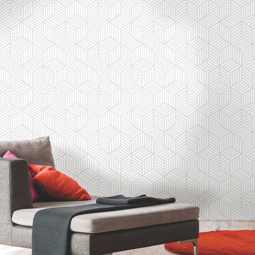 A modern room featuring a gray chaise lounge with orange and red pillows, a dark gray throw, and a vibrant orange circular rug. The wall is adorned with WallPops Striped Hexagon Peel and Stick Wallpaper showcasing a white geometric pattern of thin gray stripes. The overall setting is minimal and stylish.